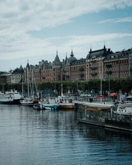 Angedockte Boote entlang einer Uferpromenade mit historischen Gebäuden im Hintergrund unter einem teilweise bewölkten Himmel.