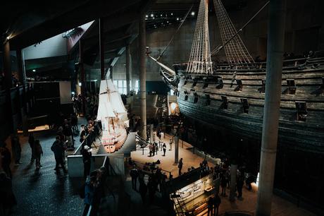 Besucher erkunden ein Museum mit großen historischen Schiffen, darunter ein erhaltenes hölzernes Kriegsschiff und ein Modellsegelboot.
