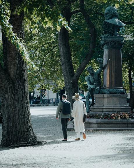 Zwei Personen gehen einen von Bäumen gesäumten Weg in der Nähe einer großen Statue in einem sonnenbeschienenen Park entlang.
