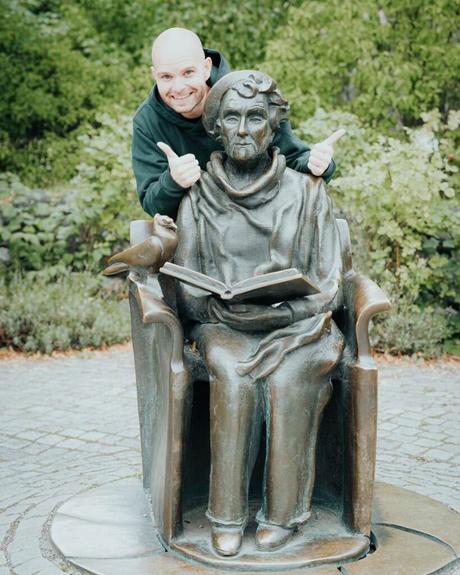 Ein lächelnder Mann zeigt den Daumen nach oben hinter einer Bronzestatue einer sitzenden Frau, die im Freien ein Buch liest.