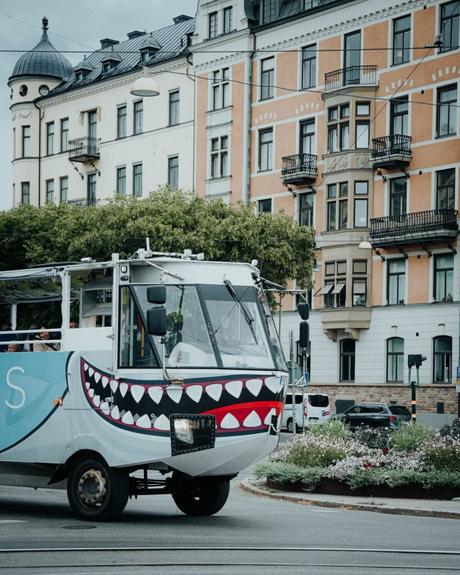 Ein weißer, amphibischer Tourbus mit aufgemalten Haifischzähnen fährt an verschnörkelten Gebäuden in einer Stadt vorbei.