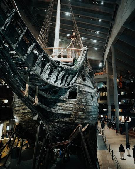 Ein großes, altes Holzschiff, das in einem Innenraum ausgestellt ist und auf dem die Menschen in einer Museumsumgebung herumlaufen.