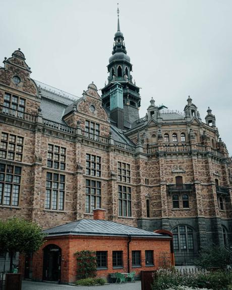 Großes historisches Backsteingebäude mit verschnörkelten Türmen hinter einem kleinen roten Backsteinhaus und Grünanlagen.