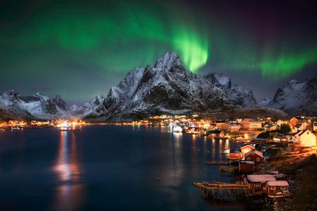 Nordlichter leuchten über verschneiten Bergen und einem nachts beleuchteten Küstendorf.