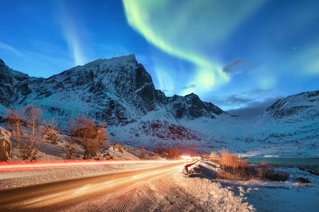 Nordlichter leuchten über schneebedeckten Bergen und einer kurvenreichen Straße mit roten und weißen Autolichtspuren.