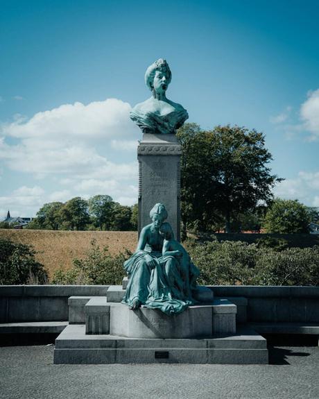 Bronzestatue einer sitzenden Frau mit einer Büste einer anderen Frau auf einem Sockel hinter ihr, im Freien.