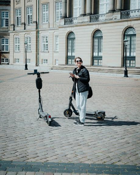 Person, die mit zwei Elektrorollern auf einer Kopfsteinpflasterstraße steht, historisches Gebäude im Hintergrund.