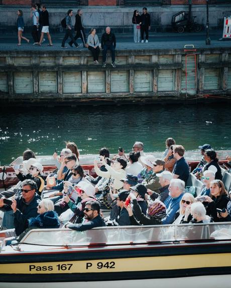 Touristen sitzen auf einer Bootstour in einem Kanal, während im Hintergrund Menschen am Ufer entlang spazieren und sitzen.
