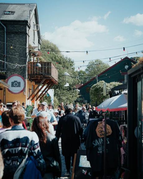 Eine belebte Marktszene im Freien mit Menschen, die an Geschäften vorbeilaufen, und Lichterketten über dem Kopf.