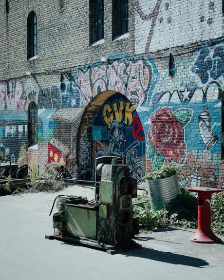 Eine verrostete grüne Maschine steht an einem sonnigen Tag vor einer mit Graffiti beschmierten Backsteinmauer.