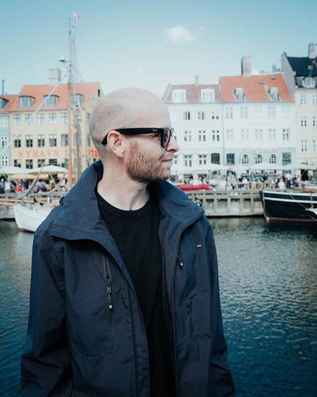 Ein Mann mit Sonnenbrille und Jacke steht an einem Kanal mit bunten Gebäuden und Booten im Hintergrund.