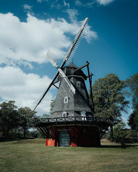 Eine traditionelle Windmühle steht auf einer Wiese unter einem teilweise bewölkten blauen Himmel, umgeben von Bäumen.