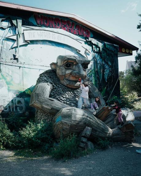 Drei Kinder klettern auf eine große hölzerne Troll-Skulptur vor einem Wandgemälde mit der Aufschrift Wunderland.