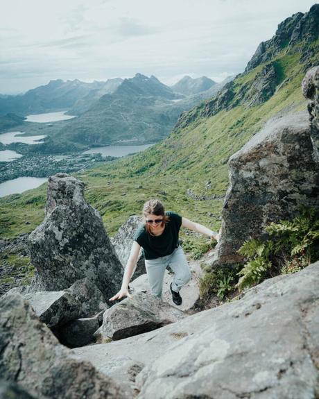 Eine Frau wandert auf einem felsigen Bergpfad mit grünen Hügeln und Seen im Hintergrund.