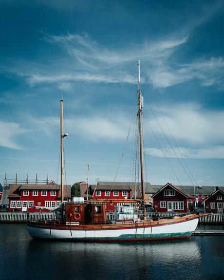 Ein weißes Segelboot liegt auf dem ruhigen Wasser vor roten Holzhäusern unter blauem Himmel.