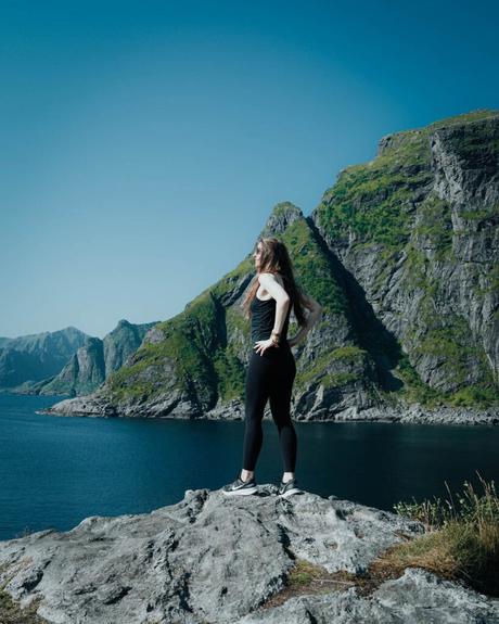 Frau in Sportkleidung steht auf einem Felsen mit Blick auf einen blauen See und grüne Berge unter einem klaren Himmel.