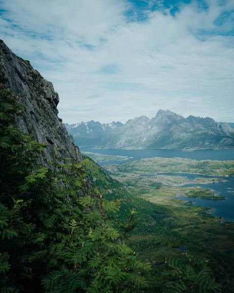 Berglandschaft mit Felsen, grünem Tal, Wasser und fernen Gipfeln unter einem teilweise bewölkten Himmel.