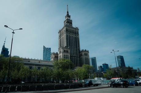 Hohes historisches Gebäude mit Turmspitze, umgeben von Bäumen und modernen Wolkenkratzern unter blauem Himmel.