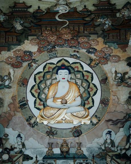 Wandgemälde mit einem sitzenden Buddha im Lotussitz, umgeben von floralen und himmlischen Figuren.