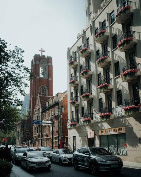 Straßenansicht mit Autos, einer Kirche mit einem roten Kreuz und einem Gebäude mit blumengeschmückten Balkonen.