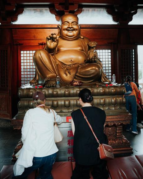 Zwei Menschen knien und beten vor einer großen goldenen lachenden Buddha-Statue in einem Tempel.