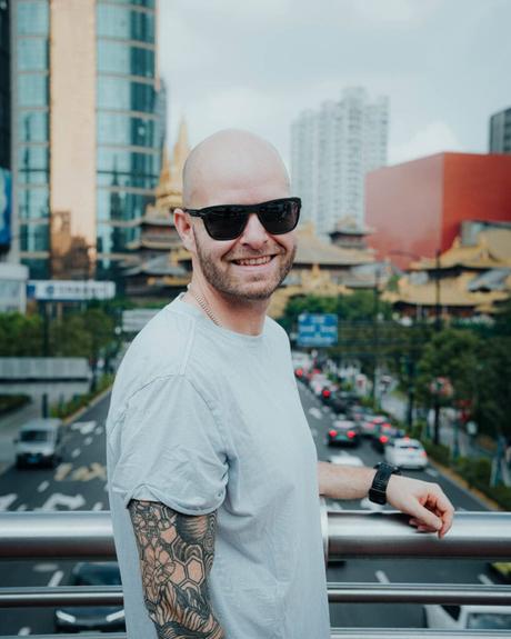 Ein lächelnder Mann mit Sonnenbrille und tätowiertem Arm steht auf einer Stadtbrücke mit Verkehr und Gebäuden im Hintergrund.