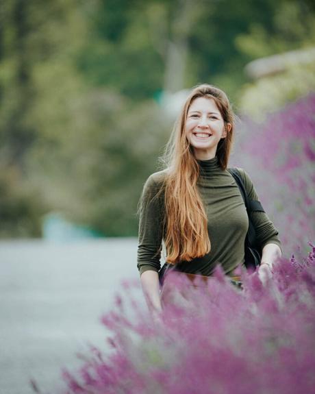 Eine lächelnde Frau mit langen Haaren steht im Freien neben leuchtend violetten Blumen und Grünpflanzen.