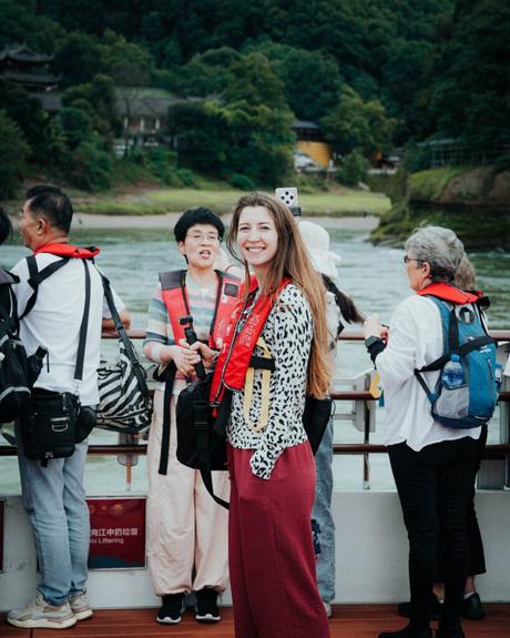 Eine lächelnde Frau in einer roten Schwimmweste steht auf einem Boot mit anderen Menschen, mit grünen Hügeln im Hintergrund.
