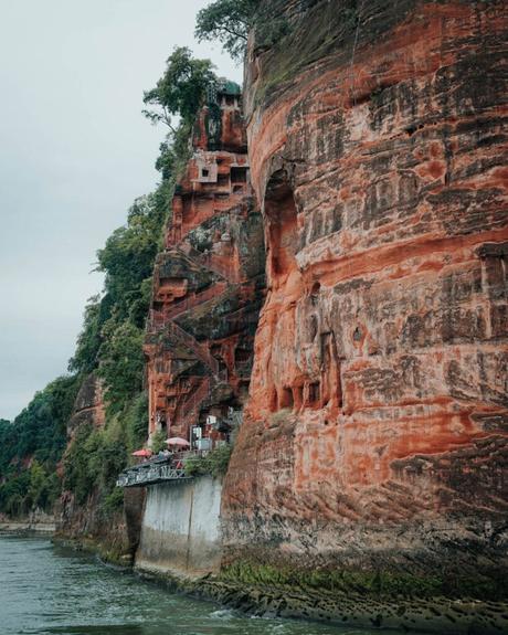 Steile rote Felsen mit geschnitzten Stufen und Grün, neben einem Fluss unter einem bewölkten Himmel.