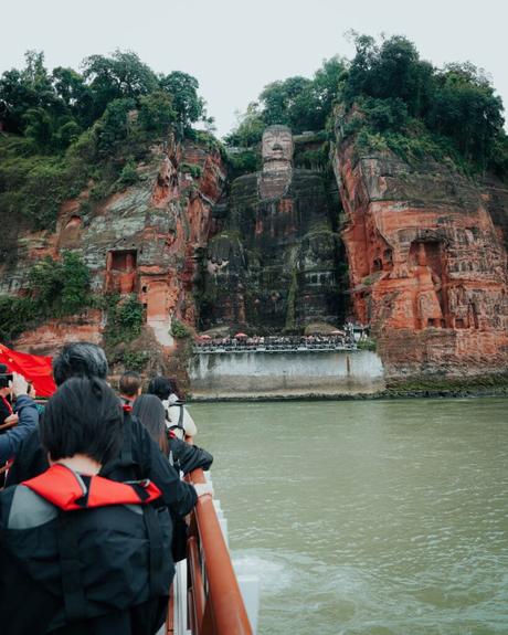 Menschen auf einem Boot betrachten den Riesenbuddha von Leshan, der in einen roten Felsen am Fluss gehauen und von Grün umgeben ist.