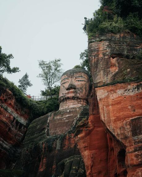 Riesige Buddha-Statue aus Stein, die in einen roten Felsen gehauen wurde, umgeben von Grün unter einem bewölkten Himmel.