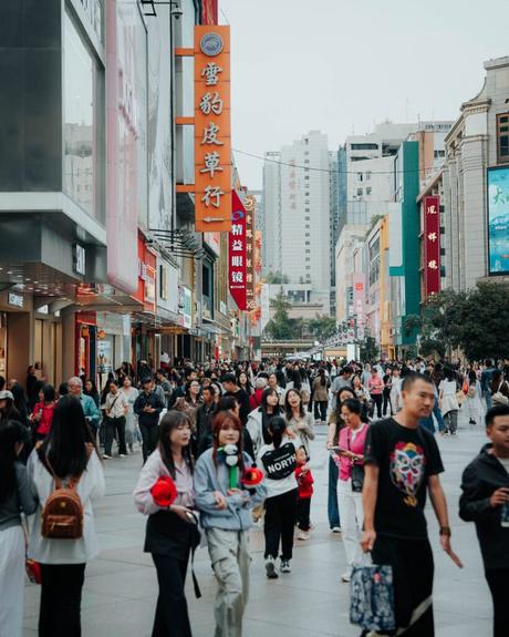 Eine belebte Stadtstraße mit Menschen, die spazieren gehen und einkaufen, umgeben von hohen Gebäuden und bunten Schildern in chinesischer Sprache.