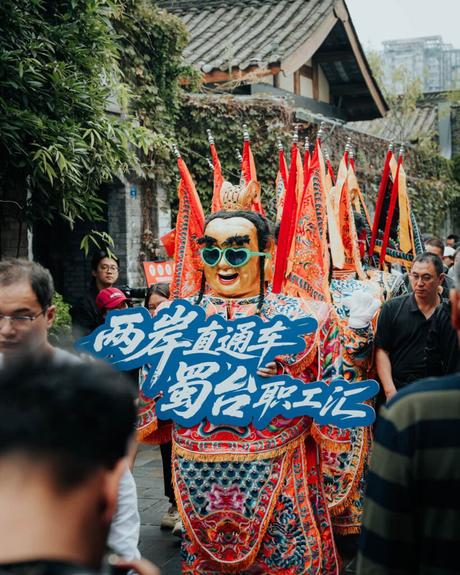 Bunte Parade mit maskierten Darstellern in kunstvollen Kostümen, die ein Schild mit chinesischen Schriftzeichen halten, auf einer belebten Straße.