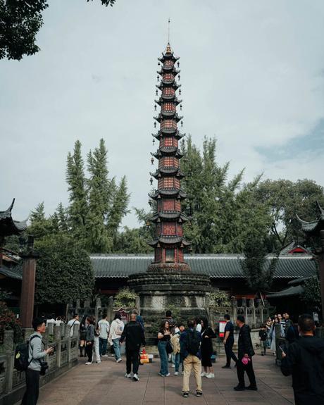 Eine hohe, verschnörkelte Pagode steht in einem Innenhof, in dem Menschen spazieren gehen und Bäume im Hintergrund zu sehen sind.