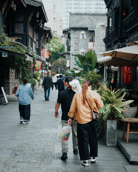 Ein Mann mit einem weißen Hund auf der Schulter geht mit einer Frau durch eine belebte, enge Straße mit Geschäften und Pflanzen.