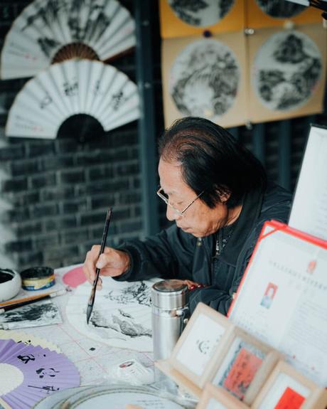 Ein Künstler malt an einem Tisch verschlungene Muster auf einen Fächer, während hinter ihm Fächer und Kunstwerke ausgestellt sind.