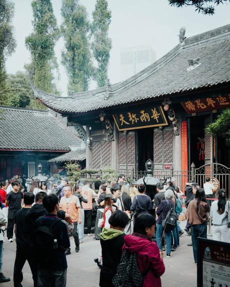 Menschenmenge beim Besuch eines traditionellen chinesischen Tempels, umgeben von Bäumen und kunstvoller Holzarchitektur.