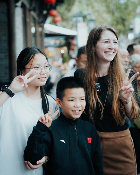 Drei lächelnde Menschen, die gemeinsam in einer belebten Gegend im Freien posieren und Friedenszeichen machen.