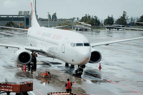 Ein weißes Passagierflugzeug auf dem nassen Rollfeld eines Flughafens mit Arbeitern und orangefarbenen Kegeln in der Nähe.