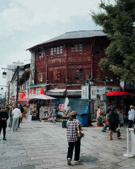 Auf einer belebten Straße in einer asiatischen Stadt gehen Menschen an Geschäften und einem großen roten Holzgebäude vorbei.