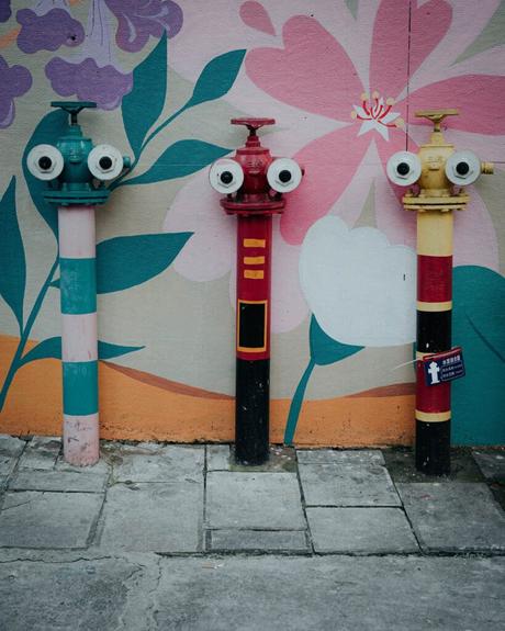 Drei farbenfrohe Hydranten mit bemalten Gesichtern stehen vor einer Blumenwand auf dem Bürgersteig.