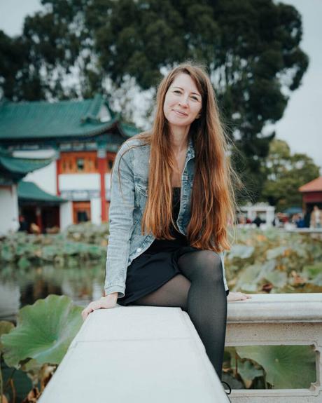 Frau mit langen Haaren und Jeansjacke sitzt lächelnd auf einer Brücke an einem Lotusteich, dahinter Bäume und Gebäude.