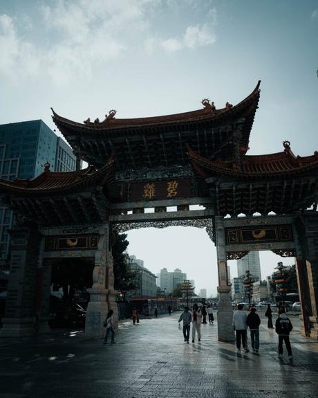 Traditionelles chinesisches Tor mit verschnörkeltem Dach, darunter gehende Menschen, Stadtgebäude im Hintergrund.