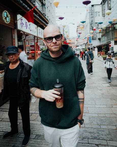 Ein Mann mit Sonnenbrille und einem Getränk steht auf einer belebten Straße mit Laternen und Geschäften im Hintergrund.