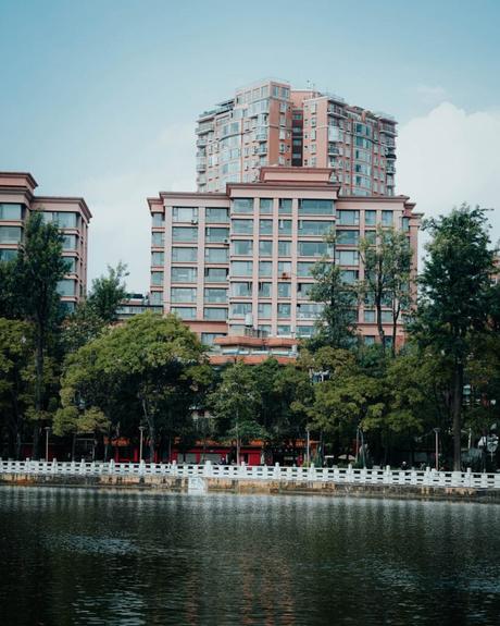 Hohe moderne Wohnhäuser mit Glasfenstern hinter Bäumen und einem See mit einem weißen Zaun davor.