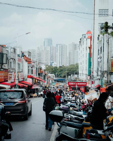 Belebte städtische Straße in China mit Menschen, Motorrollern, Geschäften und hohen Gebäuden im Hintergrund.