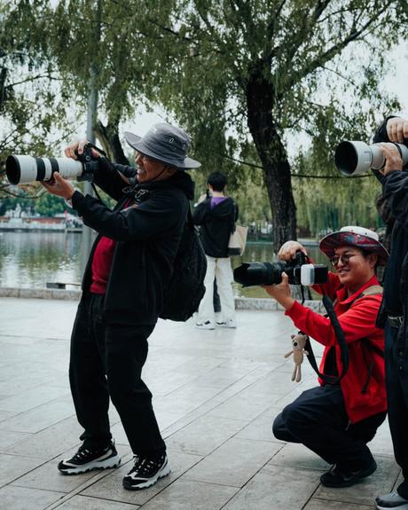 Zwei Personen mit Kameras und großen Objektiven fotografieren im Freien in der Nähe eines Flusses; Bäume und andere Menschen im Hintergrund.
