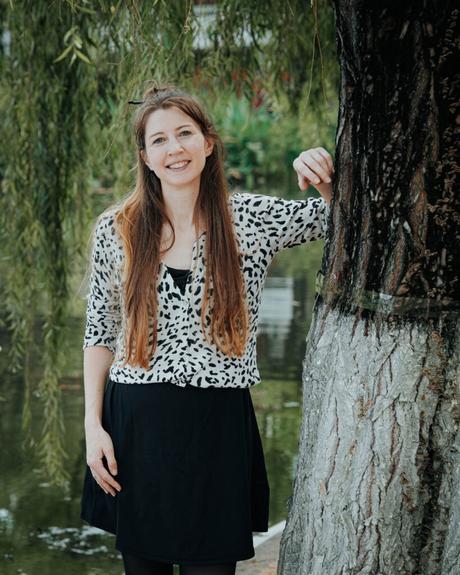 Eine lächelnde Frau mit langen Haaren steht an einem Baum in der Nähe eines Teiches und trägt ein schwarz-weißes Outfit.