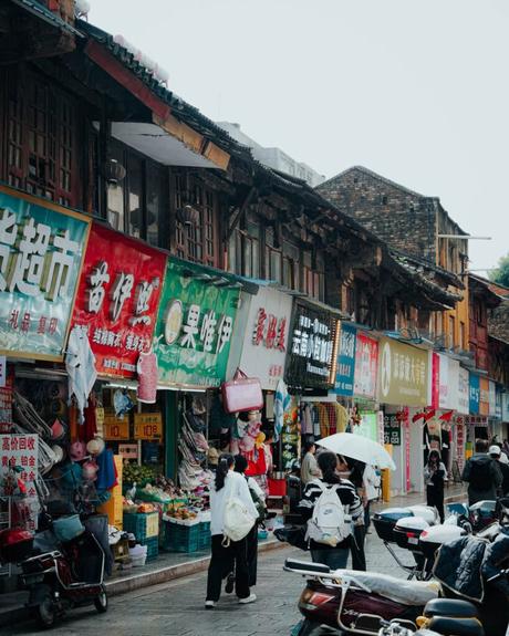 Belebter Straßenmarkt in China mit bunten Ladenschildern, Einkäufern und vor den Geschäften geparkten Motorrollern.