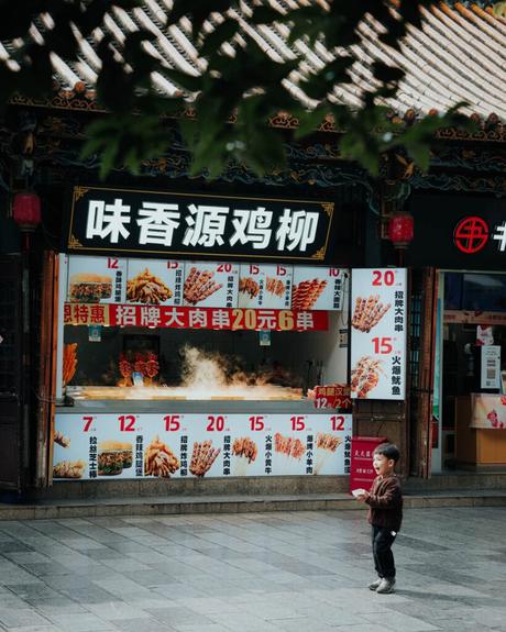 Ein kleines Kind steht vor einem Straßenimbiss mit Spießen und Schildern, auf denen Speisekarten in chinesischer Sprache abgebildet sind.